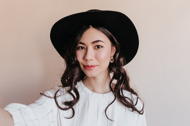 免费Photo 卷发的中国女性自拍。戴着帽子在米色背景上摆姿势的亚洲模特正面照。